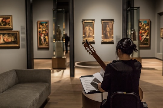 A person sits in an art gallery or museum, playing a guitar while facing a music stand with sheet music. The room has several paintings on the walls and sculptures inside glass cases. The lighting is focused on the artwork, highlighting the details and creating a cozy atmosphere.