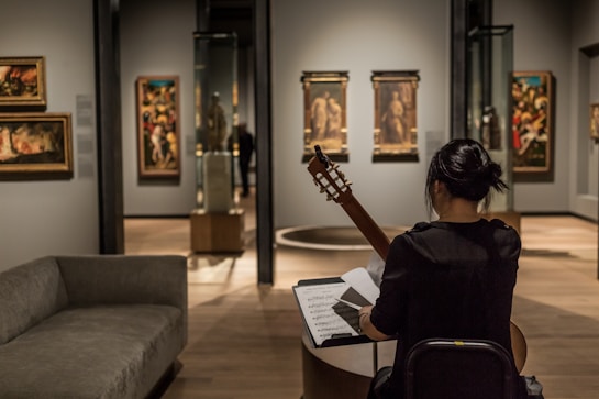A person sits in an art gallery or museum, playing a guitar while facing a music stand with sheet music. The room has several paintings on the walls and sculptures inside glass cases. The lighting is focused on the artwork, highlighting the details and creating a cozy atmosphere.