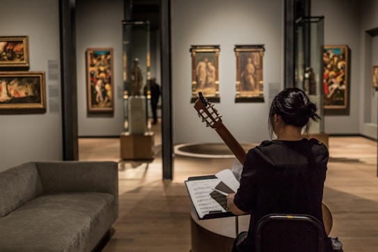 A person sits in an art gallery or museum, playing a guitar while facing a music stand with sheet music. The room has several paintings on the walls and sculptures inside glass cases. The lighting is focused on the artwork, highlighting the details and creating a cozy atmosphere.
