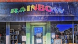 A storefront with a colorful 'RAINBOW' sign above a blue door numbered 127 is adorned with various posters. The window display includes books and other items visible inside the store.