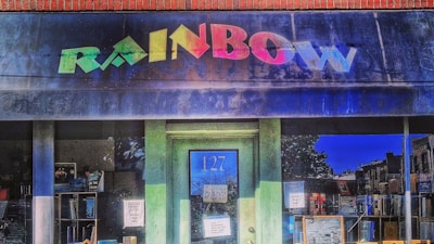 A storefront with a colorful 'RAINBOW' sign above a blue door numbered 127 is adorned with various posters. The window display includes books and other items visible inside the store.