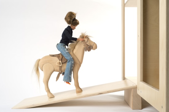 A doll with brown hair wearing a dark top and blue jeans is riding a wooden toy horse up a wooden ramp. The setting appears minimalistic with a plain background, and the horse is detailed with saddle and bridle.