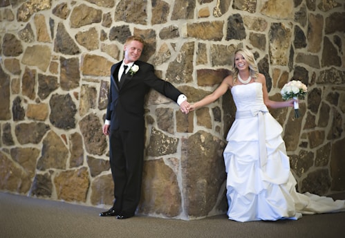 A couple in wedding attire is standing against a stone wall. The groom, in a black suit with a white boutonniere, is smiling and leaning against the wall. The bride, wearing a white gown and holding a bouquet of white flowers, is smiling and leaning towards the groom with outstretched arms.