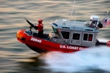 A U.S. Coast Guard boat is speeding through water with a crew member in a bright red suit operating a mounted weapon. The boat is marked with the number 25640 and has an American flag and equipment on it.
