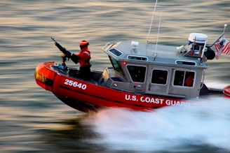 A U.S. Coast Guard boat is speeding through water with a crew member in a bright red suit operating a mounted weapon. The boat is marked with the number 25640 and has an American flag and equipment on it.