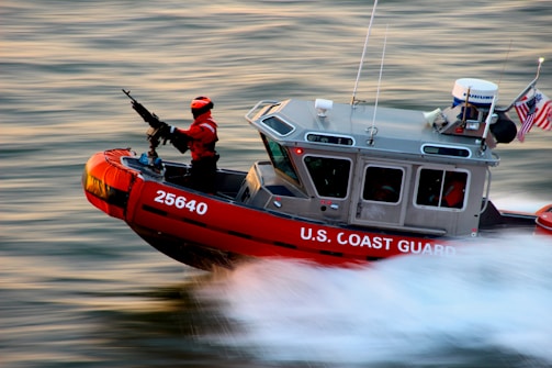 A U.S. Coast Guard boat is speeding through water with a crew member in a bright red suit operating a mounted weapon. The boat is marked with the number 25640 and has an American flag and equipment on it.