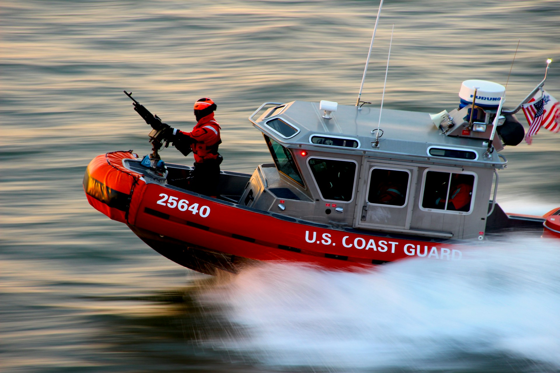 A U.S. Coast Guard boat is speeding through water with a crew member in a bright red suit operating a mounted weapon. The boat is marked with the number 25640 and has an American flag and equipment on it.