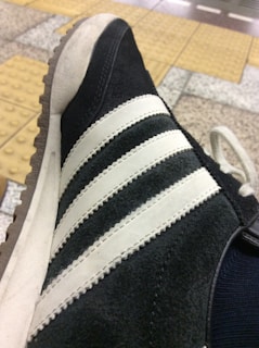A close-up view of a sneaker with black suede material and white leather stripes, featuring a gum outsole. The background consists of a tiled floor with yellow tactile paving.