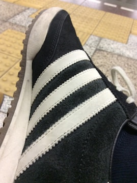 A close-up view of a sneaker with black suede material and white leather stripes, featuring a gum outsole. The background consists of a tiled floor with yellow tactile paving.
