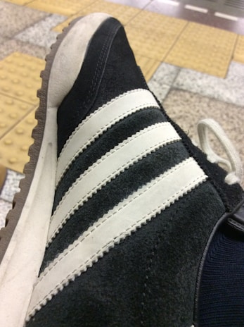 A close-up view of a sneaker with black suede material and white leather stripes, featuring a gum outsole. The background consists of a tiled floor with yellow tactile paving.