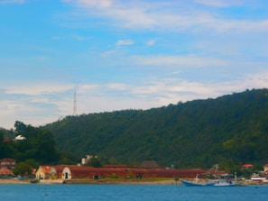 A coastal scene featuring a calm blue sea in the foreground with two boats anchored near a shoreline dotted with houses with red roofs. Green hills rise in the background under a partly cloudy sky. A communication tower is visible on the hilltop.