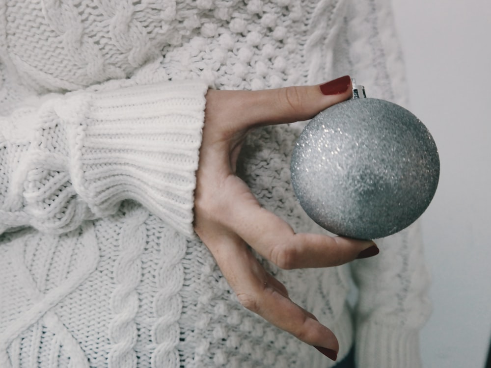 A person wearing a knitted sweater holding a silver Christmas ornament.
