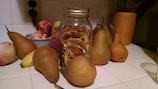 A glass jar filled with dried fruits is surrounded by fresh pears, apples, and a banana on a white tiled kitchen countertop. There's a wooden utensil holder and bowl in the background.