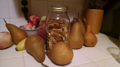 A glass jar filled with dried fruits is surrounded by fresh pears, apples, and a banana on a white tiled kitchen countertop. There's a wooden utensil holder and bowl in the background.