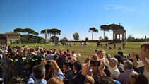A large crowd of people is gathered in a grassy area with a few trees and a small building in the background. Several people are taking photos with their phones. In the distance, a group of horses and handlers are present, along with a flock of sheep. The bright blue sky and sunlight indicate a clear day.