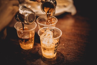 Three transparent plastic cups on a wooden surface being filled with a caramel-colored liquid from metal containers. The first cup contains ice cubes, and the background is slightly blurred.