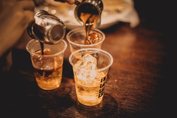 Three transparent plastic cups on a wooden surface being filled with a caramel-colored liquid from metal containers. The first cup contains ice cubes, and the background is slightly blurred.