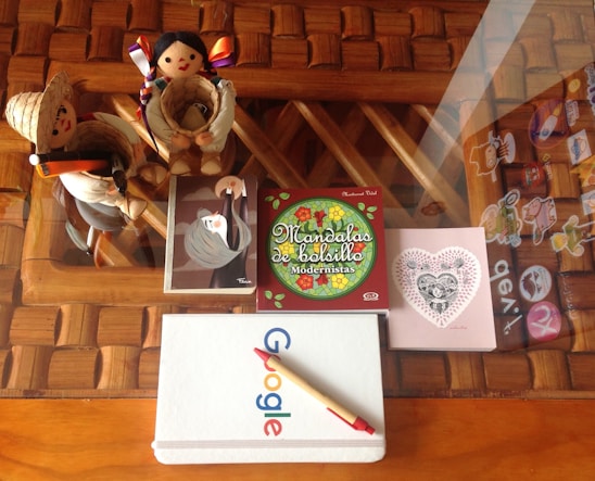 A wooden table with a woven design holds a variety of items. There are two plush dolls, one dressed in a traditional outfit with a straw hat and the other with colorful ribbons in its hair. Beside these dolls, a notebook with the Google logo rests on the table, with a pen laying across it. Also present is a small colorful book titled 'Mandalas de bolsillo Modernistas' with floral art on the cover. Accompanying these is a small card with heart and floral motifs featuring a woman's face. The glass surface of the table displays decorative stickers and reflections, adding a whimsical touch.