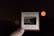 A hand holds a vintage Kodak Kodachrome slide against a dark background. The slide contains an image of a serene lake scene at night, with a faint shoreline and hills in the distance. A reflection of the landscape is visible on the water surface.