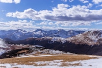 A breathtaking mountainous landscape with snow-covered peaks under a partly cloudy sky. The foreground features a mix of grass and patches of snow, leading into rolling hills and dense forests. The background showcases jagged mountain ranges illuminated by sunlight, creating a majestic and serene atmosphere.