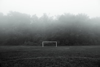 A football goalpost is centered on a foggy field, surrounded by dense trees and fog that obscure the background, creating a mysterious atmosphere.