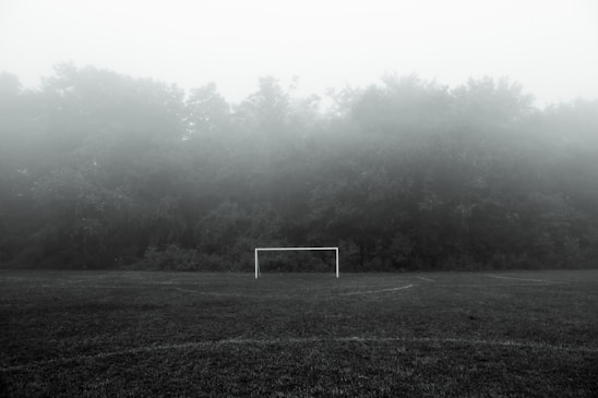 A football goalpost is centered on a foggy field, surrounded by dense trees and fog that obscure the background, creating a mysterious atmosphere.
