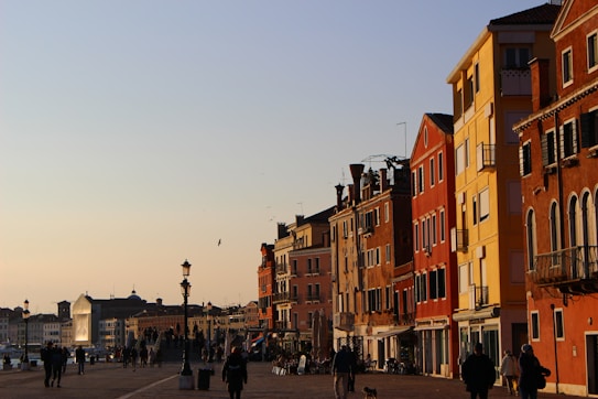 A scenic view of a city street lined with vibrant, colorful buildings in hues of red, yellow, and orange. The street is bustling with people walking, and a few pedestrians are silhouetted against the setting sun. The architecture is reminiscent of historic European styles, with the buildings featuring arched windows and intricate detailing. A lamp post adds to the nostalgic atmosphere, and the sky above is a clear blue, transitioning to a light orange near the horizon.