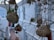 A collection of woven bird nests hangs from thin branches, set against a backdrop of white buildings. The scene includes some red flowers and greenery. The nests have an intricate design and are made of straw-like materials, creating an organic feel within the urban setting.
