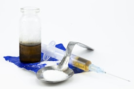 A syringe with a needle, a small glass vial containing a brown liquid, a metal spoon with white powder, and a piece of torn blue plastic are arranged against a white background.