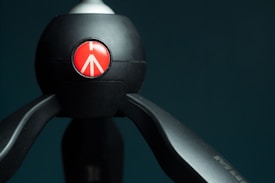 A black tripod head with a sleek design, featuring a prominent red circular emblem with a white arrow-like symbol in the center. The background is a dark teal color, providing contrast to the black and red elements.