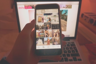A person holds a smartphone displaying a social media app with various photos. The phone is viewed in front of an open laptop, likely in a dimly lit or warm-toned room. The laptop screen displays a blurred webpage in the background.
