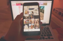 A person holds a smartphone displaying a social media app with various photos. The phone is viewed in front of an open laptop, likely in a dimly lit or warm-toned room. The laptop screen displays a blurred webpage in the background.