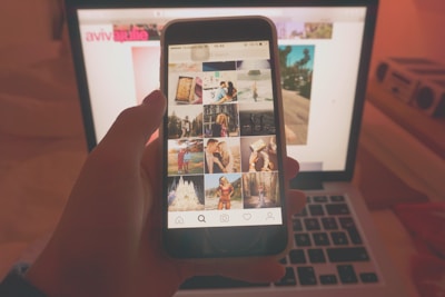 A person holds a smartphone displaying a social media app with various photos. The phone is viewed in front of an open laptop, likely in a dimly lit or warm-toned room. The laptop screen displays a blurred webpage in the background.