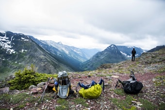 A scenic mountain landscape with a person standing on a rocky outcrop gazing at the expansive valley and mountain ranges in the distance. Three backpacks and hiking gear are scattered in the foreground on the grassy and rocky terrain. Snow-capped peaks and dense clouds dominate the background, adding depth and a sense of vastness to the scene.