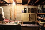 A dimly lit basement or utility room with concrete block walls and exposed ceiling beams. The room has a utility sink with various cleaning supplies and tools around it, including a drying rack, a mop, and a bucket. Above the sink and to the right is a small cabinet with metallic containers on top. To the right, there are shelves filled with jars and canned goods stored in boxes. A single light bulb hangs from the ceiling, illuminating the space.