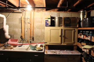 A dimly lit basement or utility room with concrete block walls and exposed ceiling beams. The room has a utility sink with various cleaning supplies and tools around it, including a drying rack, a mop, and a bucket. Above the sink and to the right is a small cabinet with metallic containers on top. To the right, there are shelves filled with jars and canned goods stored in boxes. A single light bulb hangs from the ceiling, illuminating the space.