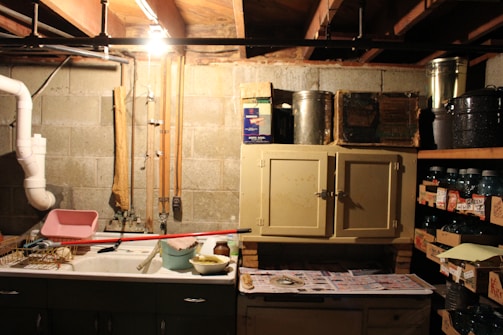 A dimly lit basement or utility room with concrete block walls and exposed ceiling beams. The room has a utility sink with various cleaning supplies and tools around it, including a drying rack, a mop, and a bucket. Above the sink and to the right is a small cabinet with metallic containers on top. To the right, there are shelves filled with jars and canned goods stored in boxes. A single light bulb hangs from the ceiling, illuminating the space.