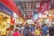 A bustling indoor market filled with people shopping and browsing various stalls that sell clothing, souvenirs, and food. The market is vibrant with colorful signage, decorations, and displays of merchandise. The scene is lively and crowded, conveying a sense of a busy shopping environment.