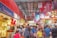 A bustling indoor market filled with people shopping and browsing various stalls that sell clothing, souvenirs, and food. The market is vibrant with colorful signage, decorations, and displays of merchandise. The scene is lively and crowded, conveying a sense of a busy shopping environment.