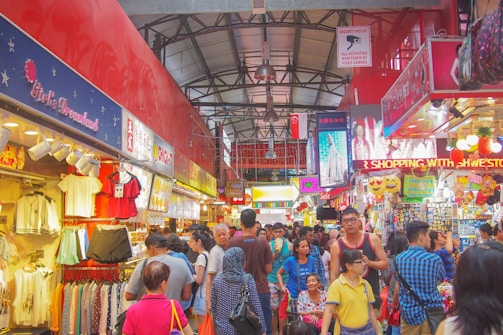 A bustling indoor market filled with people shopping and browsing various stalls that sell clothing, souvenirs, and food. The market is vibrant with colorful signage, decorations, and displays of merchandise. The scene is lively and crowded, conveying a sense of a busy shopping environment.