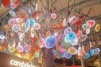 A vibrant display of large, colorful lollipops hanging from a tree-like structure. The lollipops are in various swirled patterns and colors, creating a whimsical and playful atmosphere. The background shows a signage with the word 'candylicious' illuminated, emphasizing the candy theme.