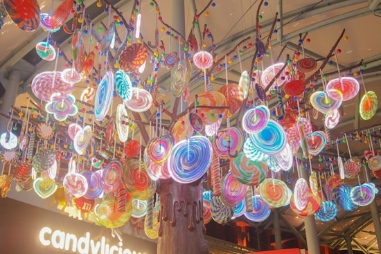 A vibrant display of large, colorful lollipops hanging from a tree-like structure. The lollipops are in various swirled patterns and colors, creating a whimsical and playful atmosphere. The background shows a signage with the word 'candylicious' illuminated, emphasizing the candy theme.
