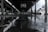 A wet, outdoor basketball court under a large, open structure with metal beams. Drops of rain are splashing onto the surface, creating ripples in the puddles. The background is slightly blurred with an overcast sky, giving a gloomy atmosphere.
