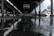 A wet, outdoor basketball court under a large, open structure with metal beams. Drops of rain are splashing onto the surface, creating ripples in the puddles. The background is slightly blurred with an overcast sky, giving a gloomy atmosphere.