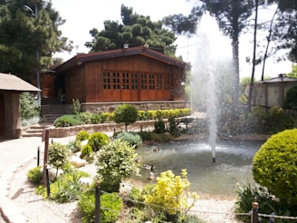 A serene garden scene features a rustic wooden house with large windows set amidst lush greenery. In the foreground, a circular pond with a fountain creates a dynamic water display surrounded by manicured shrubs and flowering plants. The sunlight filters through tall trees that border the garden, casting soft shadows.