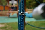 Close-up view of a blue rope held together by a metal clasp, with a blurred background showing outdoor wooden play structures.