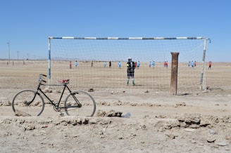 A bicycle rests on rough, dusty ground in front of a weathered soccer goal post. In the background, two teams of players in blue and red jerseys are playing football on a dry, barren field. The sky is clear and bright, suggesting a sunny day.