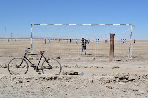 A bicycle rests on rough, dusty ground in front of a weathered soccer goal post. In the background, two teams of players in blue and red jerseys are playing football on a dry, barren field. The sky is clear and bright, suggesting a sunny day.