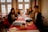 A group of young people are seated around a long table in a warmly lit dining area. They are smiling and appear to be ready to order, holding menus and surrounded by tableware. The room features large windows allowing natural light and has decorative curtains.