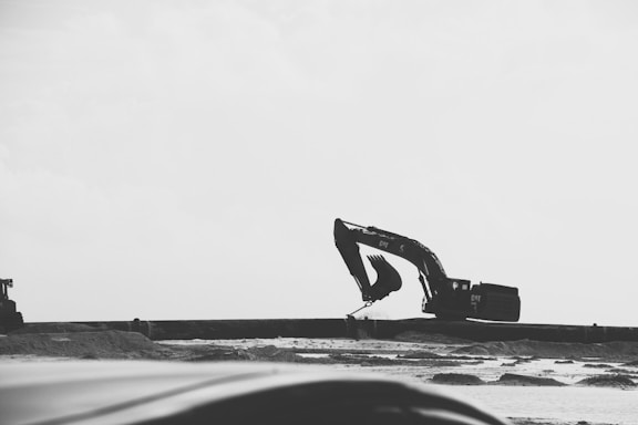 A monochrome scene with an excavator at a construction site, raising its arm against a clear sky. The background is minimalist with a focus on the machinery and sandy landscape.